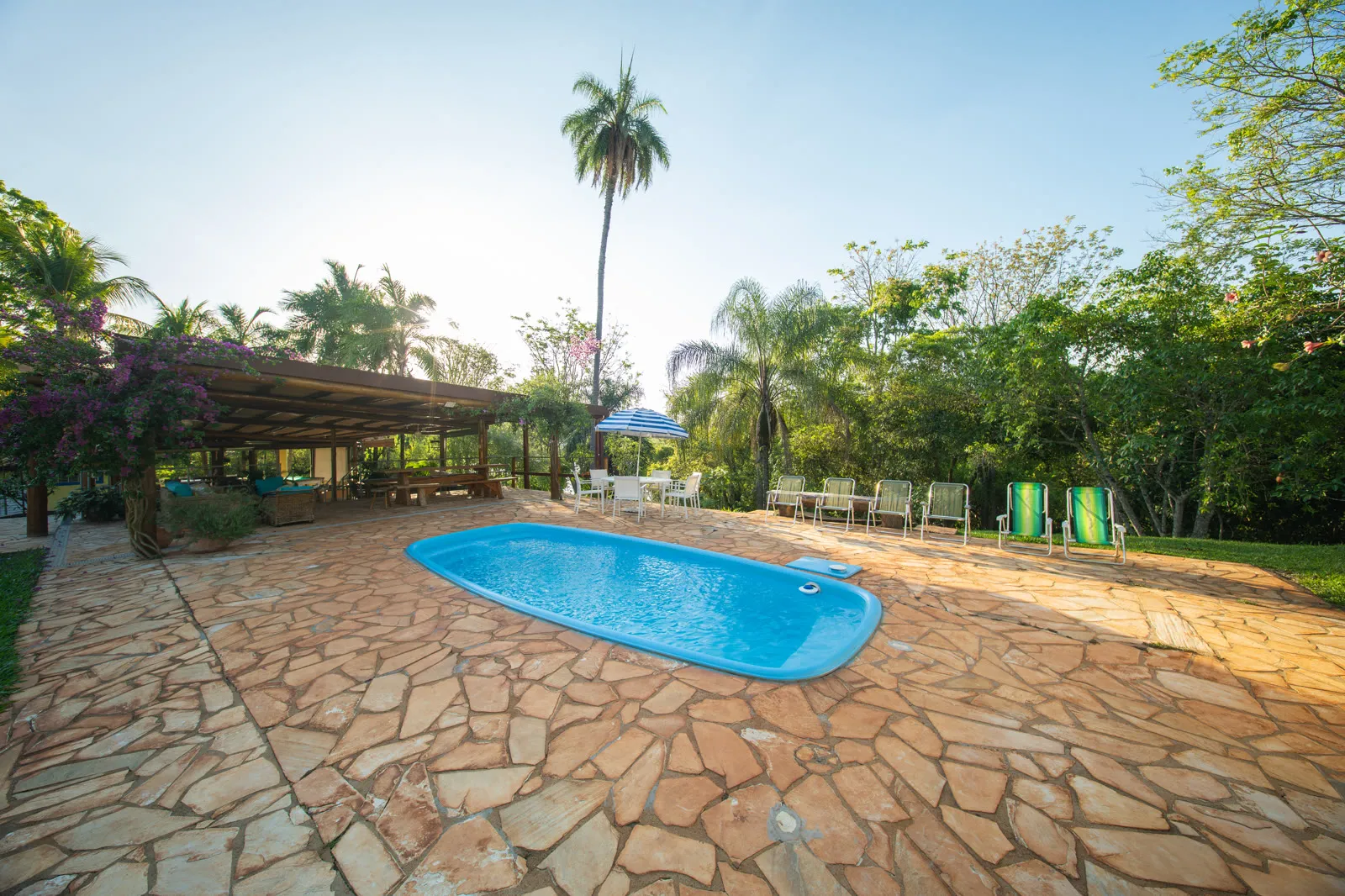 Piscina azul com deck de pedra entre coqueiros, ao lado do quiosque