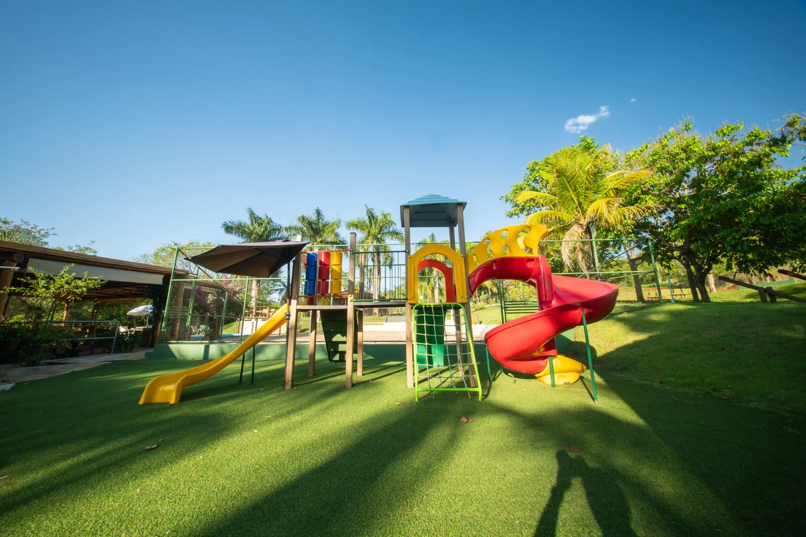 Playground colorido em grama sintética, com o quiosque ao fundo